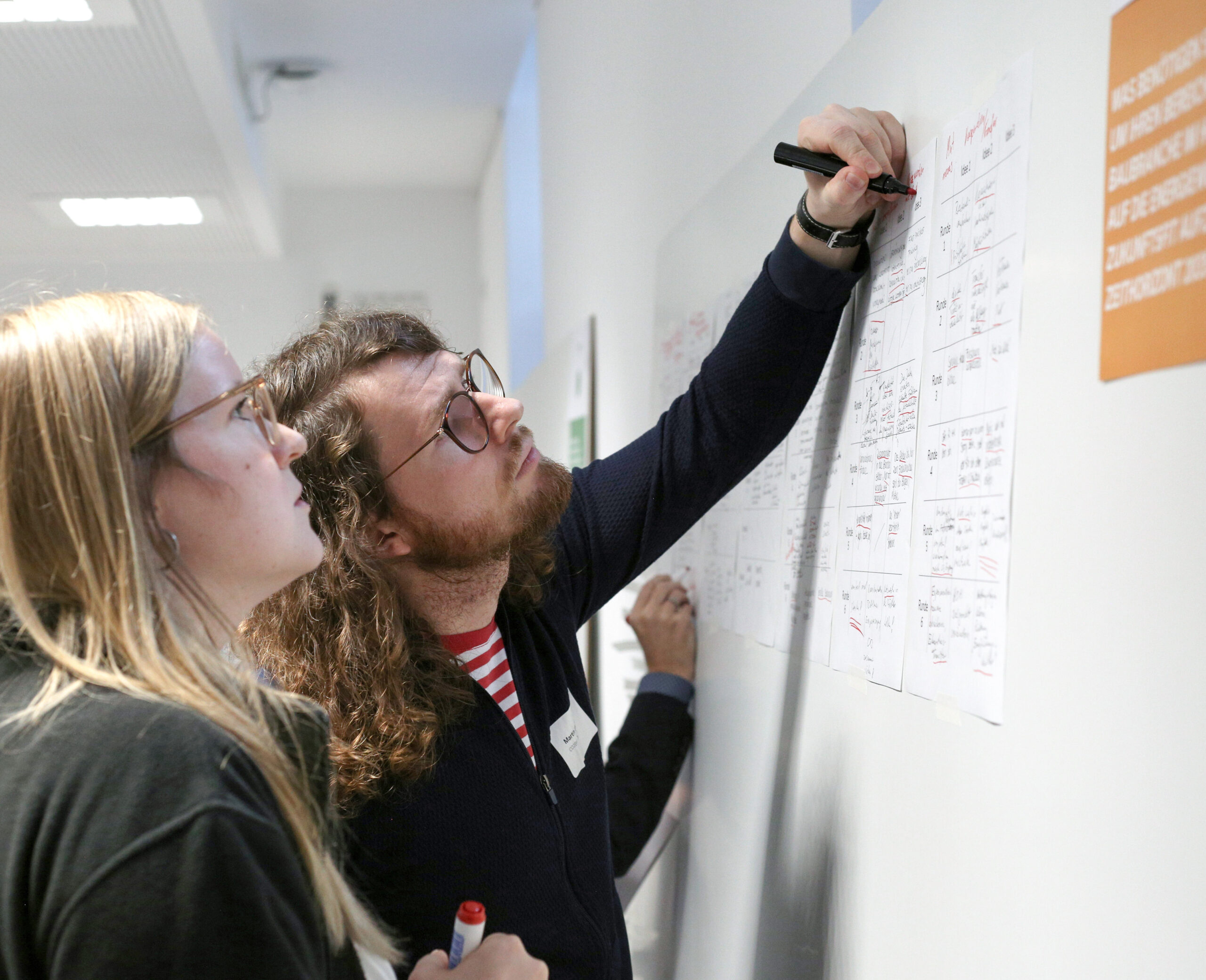 Zwei Personen die auf ein Whiteboard schauen. Eine Person schreibt etwas.